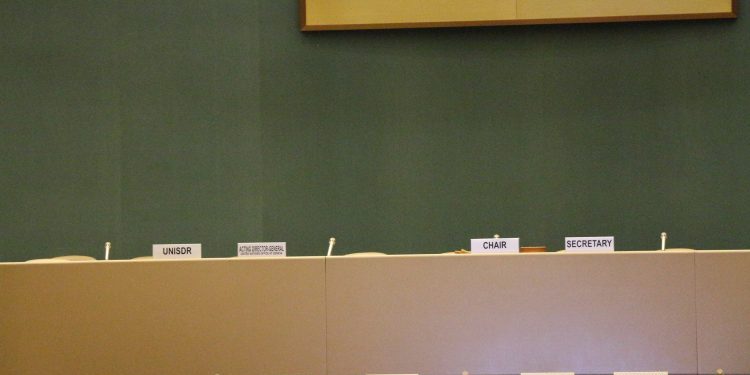The empty UN council chamber with seats and nation placards at each, a green wall behind with the bottom of the UN symbol in a frame visible above the tables