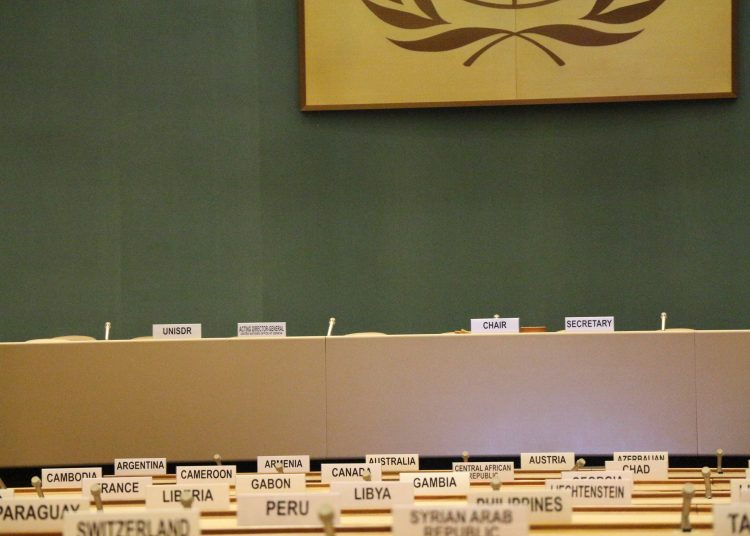 The empty UN council chamber with seats and nation placards at each, a green wall behind with the bottom of the UN symbol in a frame visible above the tables