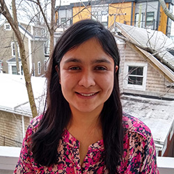 Romita Mitra – a woman with light brown skin and long dark hair looking outside with snowy city buildings behind her, looking at the camera and smiling
