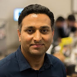 Ramesh Raskar - a medium brown skinned man with short black hair wearing a dark blue button down business shirt, looking at the camera and smiling