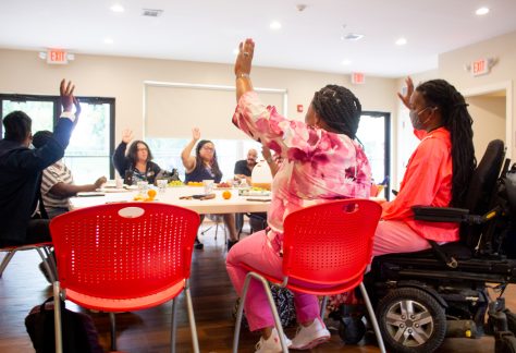 A group of people - most of whom are Black or African American - sitting around a table in red chairs with their hands raised.