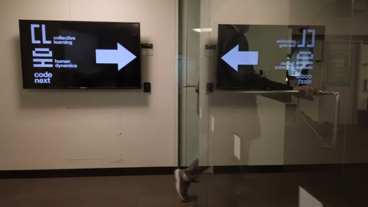 Image of a hallway entering the MIT Human Dynamics Lab. There is an LED screen on the wall with a black background and the words "collective learning" and "human dynamics" on it in white and a large white arrow pointing to the right. On the right side of the hallway is a glass wall and door. A person going through that doorway can be faintly seen.