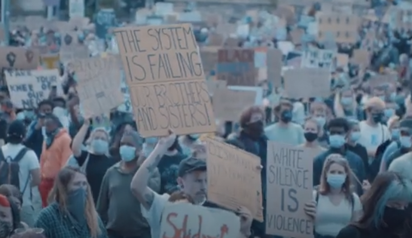 A photo of protestors holding signs at a march against racism. A person in the front holds a sign that reads "The system is failing our brothers and sisters". Next to them is another person holding a sign that says "White silence is violence"