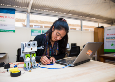 A young Latin American student with long dark hair pulled up in a ponytail is working on a robot next to an open laptop